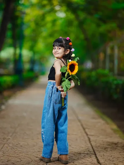A playful and shy pose, with the little girl hiding a sunflower behind her back.