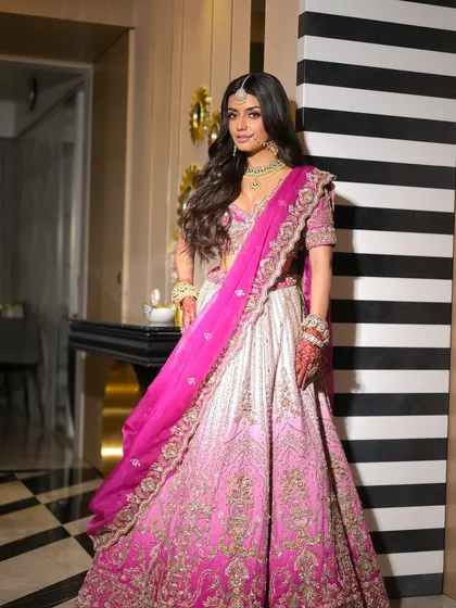 A full-length portrait against a modern backdrop. The bride looks like a contemporary princess in her pink lehenga, with makeup that enhances her natural beauty.