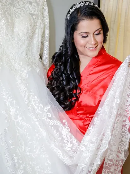 The bride-to-be in her robe, admiring the intricate lace of her wedding gown. Her hair and makeup are already done, showing the complete look before the final step.