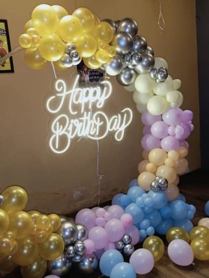 A half-arch of pastel and gold balloons with a 'Happy Birthday' neon sign, creating a modern and chic photo corner.
