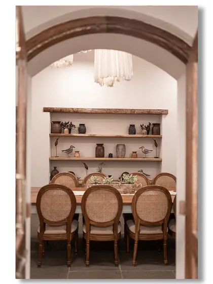 A peek through an arched doorway into a rustic dining room in a Goan villa. The long wooden table, mismatched chairs, and shelves of pottery create a warm, communal, and farmhouse-style atmosphere.
