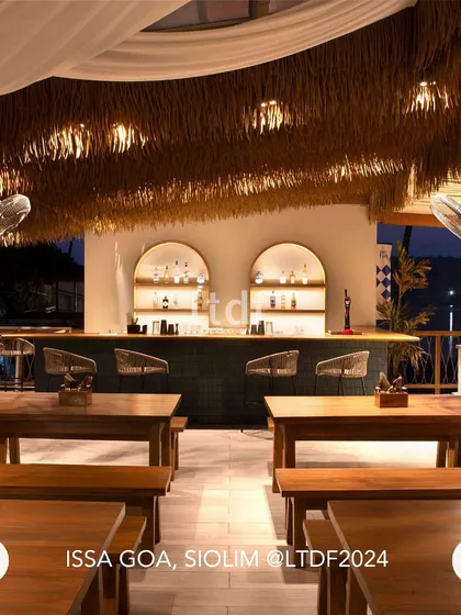 The bar on the upper deck of ISSA Goa combines a rustic thatched roof with modern, arched back-bar shelving. This blend of styles creates a sophisticated yet casual spot for enjoying a drink while taking in the panoramic waterfront views.
