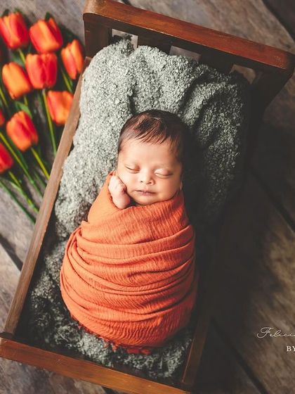 Blooming with love. This newborn is nestled in a tiny wooden bed, swaddled in a warm orange wrap next to fresh tulips, creating a beautiful, artistic portrait that feels like a dream.