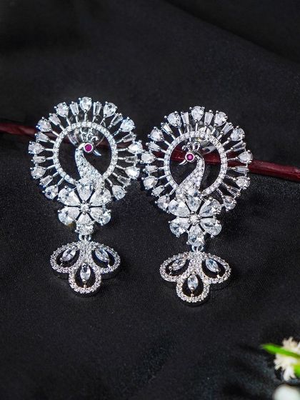 Diamond peacock earrings styled with a sprig of baby's breath on a black background. The prop adds a delicate touch without distracting from the main product.