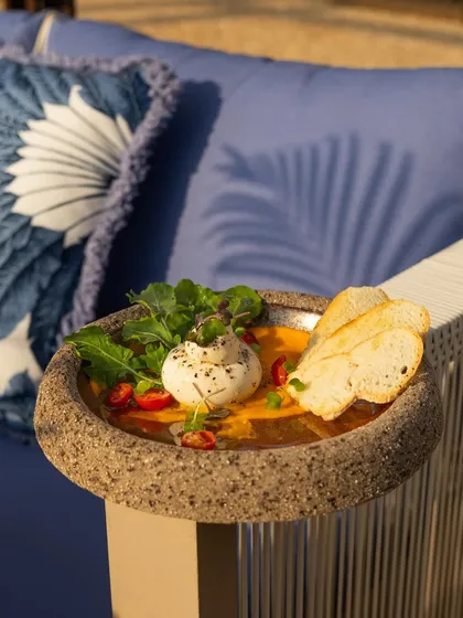 Fresh burrata served with tomato soup and crusty bread, enjoyed outdoors. The natural sunlight and casual setting make this dish look incredibly fresh and inviting.