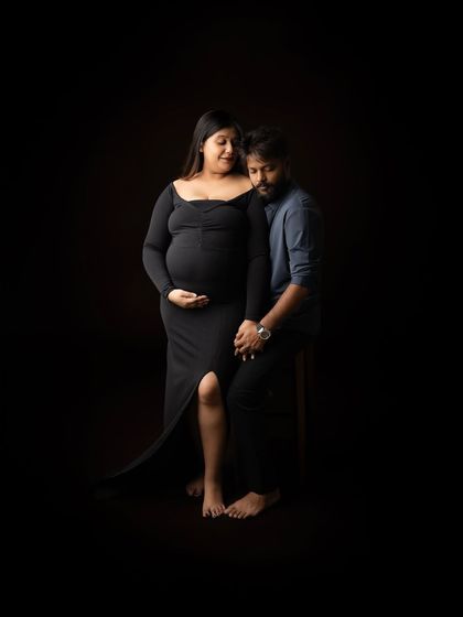 A full-length studio portrait of the couple. The mother-to-be stands in a sleek black dress while her partner sits beside her, creating a modern and stylish composition.