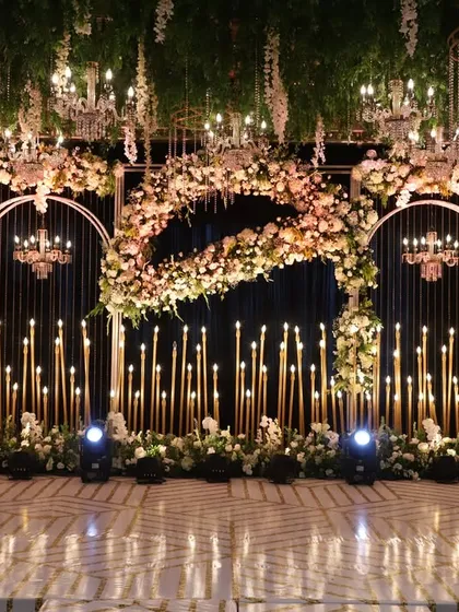 This dramatic reception stage is all about romance and light. A backdrop of golden candle-like structures is framed by arches of lush flowers, with chandeliers hanging above.