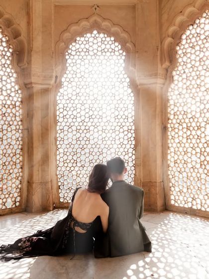 A couple finds a peaceful moment together, seated against an intricately carved 'jaali' window. The sunlight filtering through creates a breathtaking, ethereal glow, perfect for a romantic pre-wedding portrait.