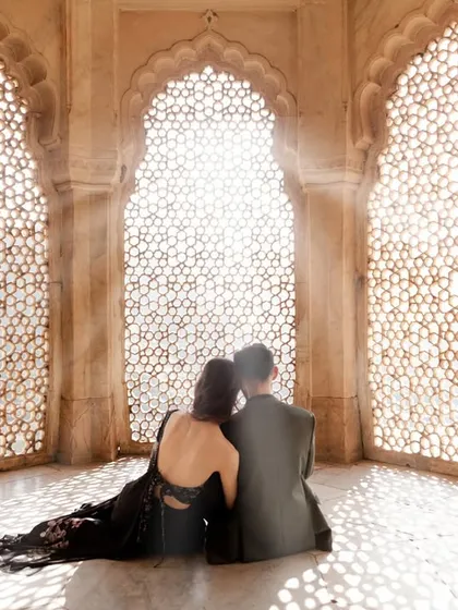 A couple finds a peaceful moment together, seated against an intricately carved 'jaali' window. The sunlight filtering through creates a breathtaking, ethereal glow, perfect for a romantic pre-wedding portrait.