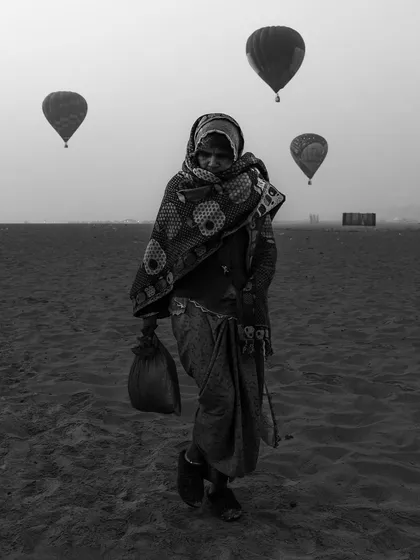 A woman walks across the sand as hot air balloons float in the sky during the Varanasi Balloon Festival, a surreal and beautiful black and white composition.