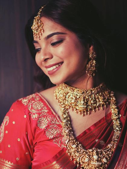 A warm, candid shot of a bride in a red saree, adorned with a heavy, traditional gold-look temple jewellery set. Her smile shows her confidence and joy.