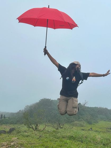 Having fun on the trail. A trekker leaps for joy with a bright red umbrella on a cloudy day at Kumara Parvatha.
