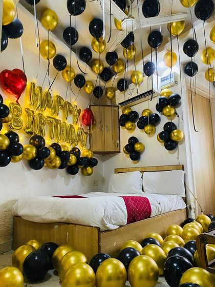 A luxurious black and gold themed birthday decoration. The room is filled with balloons on the floor and ceiling, with illuminated "Happy Birthday" foil letters above the bed.
