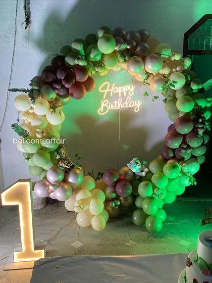 A jungle-themed first birthday party with a circular balloon arch in shades of green. The setup is enhanced with a neon 'Happy Birthday' sign and a light-up number '1'.