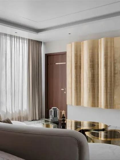 A master bedroom that is soft but makes a statement. The focal point is a sculptural, undulating gold leaf panel on the wall, which contrasts with the serene marble floors and the curved, neutral-toned sofa.