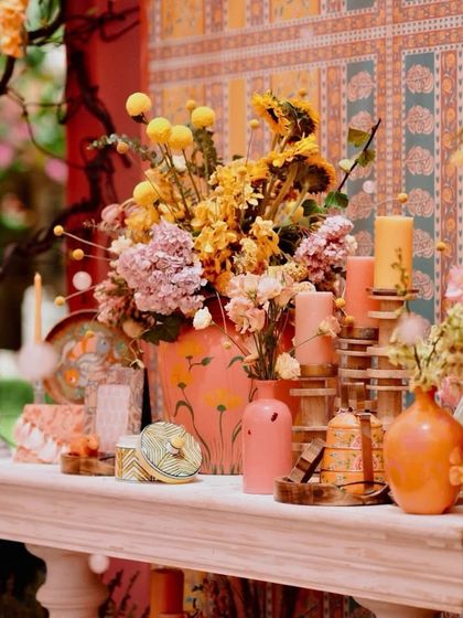A close-up of a beautifully styled tabletop at a heritage-themed haldi. The arrangement includes hand-painted vases, colorful candles, and a mix of fresh and dried flowers.