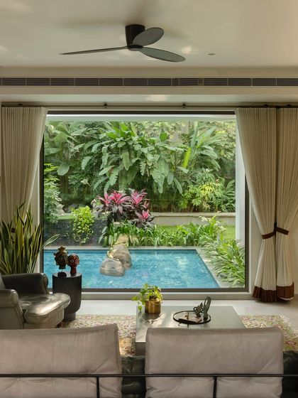 A living room view that opens directly onto a tranquil garden pool. Large glass doors and continuous flooring material blur the boundary, making the lush greenery and water feature an extension of the interior space.