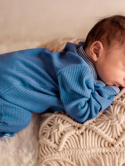 A peaceful newborn in a cozy blue outfit sleeps on their tummy on a white fur rug with a macrame pillow.