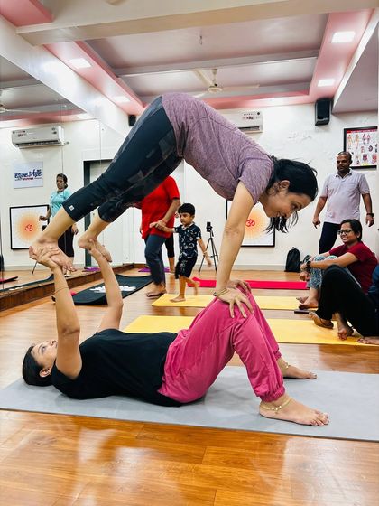 A student acts as the base while another balances in a downward dog variation. This Acro Yoga pose requires core strength and stability from both partners.