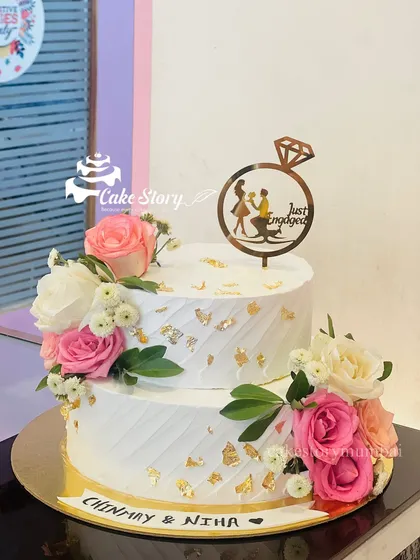 A two-tier engagement cake adorned with fresh pink and white roses. The simple textured frosting and gold leaf accents create a classic and romantic look.