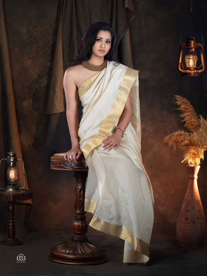 A full-length portrait showcasing a Kerala kasavu saree. The model is posed on a vintage stool amidst an earthy, rustic set, creating a timeless and artistic representation of traditional fashion.