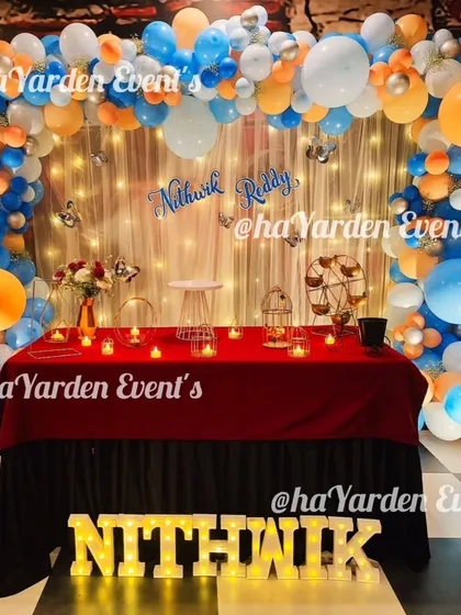 A full view of a table setup in front of a fairy light curtain, decorated with an orange and blue balloon arch.