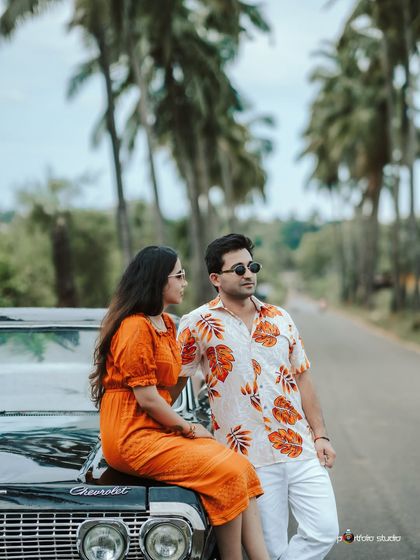 The couple poses casually on the hood of a vintage Chevrolet, creating a relaxed and stylish portrait. The palm trees and open road add to the destination feel.