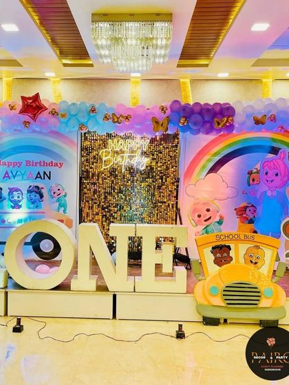 A full stage view of a Cocomelon first birthday. The setup includes a large "ONE" marquee, a school bus prop, and a backdrop featuring all the favorite characters.