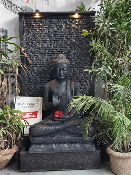 The black marble Buddha fountain is shown here surrounded by indoor plants, demonstrating its suitability for creating a serene corner within a home or office reception area.