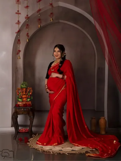 A full-length portrait showcasing a beautiful red saree with a long, flowing trail. The vintage arch and Ganesha idol create a truly traditional and blessed atmosphere.