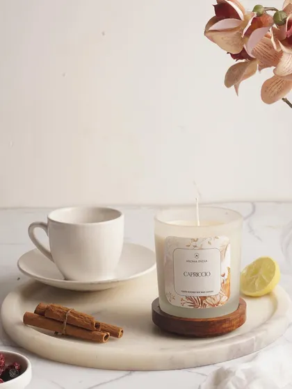 The light of a candle helps us relax and connect with our inner peace. Here, our Capriccio scented candle sits on a marble tray with cinnamon sticks, creating a serene and inspiring moment.