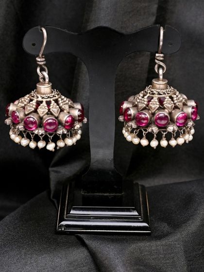 A pair of silver jhumkha earrings from a handcrafted set. They are studded with red stones and feature delicate pearl drops, showcasing traditional Indian jewelry design.
