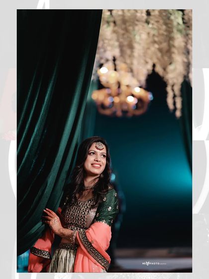 A beautiful portrait of the bride during her pre-wedding festivities. She is peeking from behind a green curtain, with a warm chandelier in the background, creating a lovely candid-style shot.