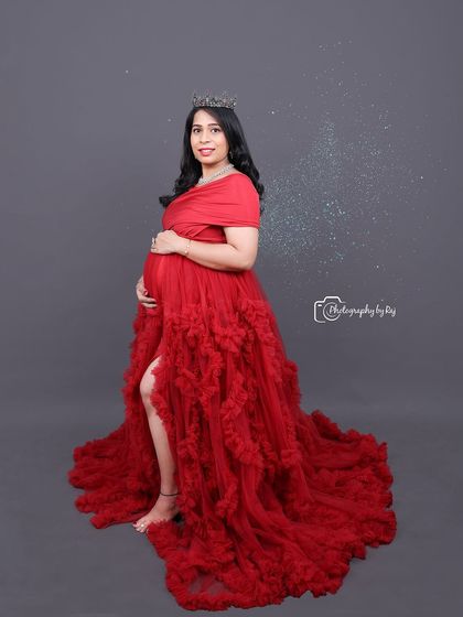 A regal studio portrait of a mother-to-be in a red ruffled gown and crown, with sparkling angel wings edited in for a magical touch.