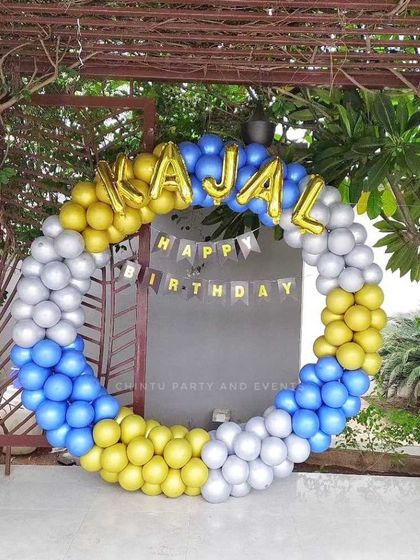 An outdoor birthday balloon ring in blue, yellow, and silver chrome. The setup includes foil letter balloons to spell out the name 'Kajal'.