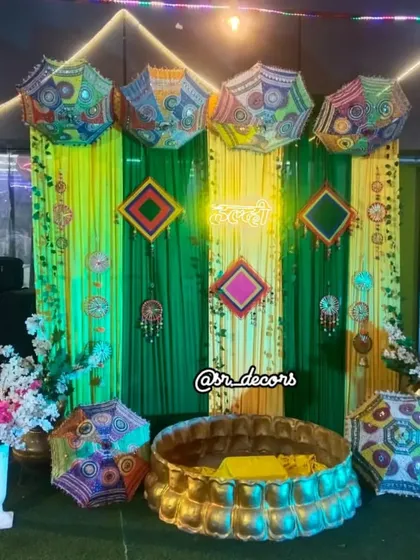 A modern Haldi setup with a twist. It includes a green backdrop, a 'Haldi' neon sign, Rajasthani umbrellas, and a traditional brass urli for the ceremony.
