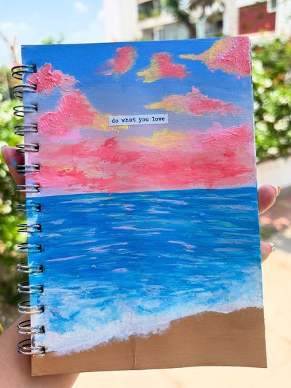 A vibrant seascape painted on the cover of my sketchbook, featuring pink clouds over a deep blue ocean. The small "do what you love" message is a personal touch.