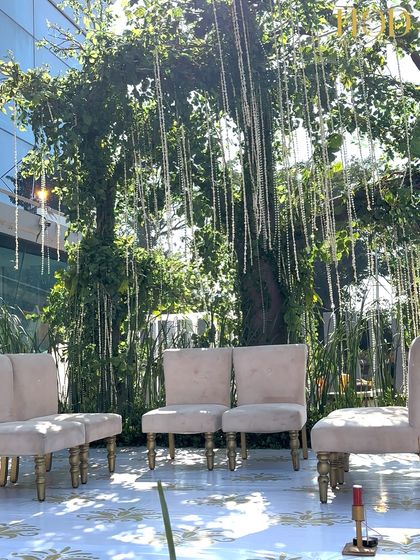 A beautiful outdoor stage for a Varmala ceremony, set against a backdrop of lush green trees with hanging pearl strings, creating a natural yet elegant look.