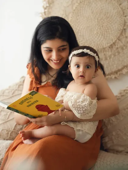 A mother reads a book to her wide-eyed baby girl. This lifestyle shot captures a quiet, everyday moment of bonding.