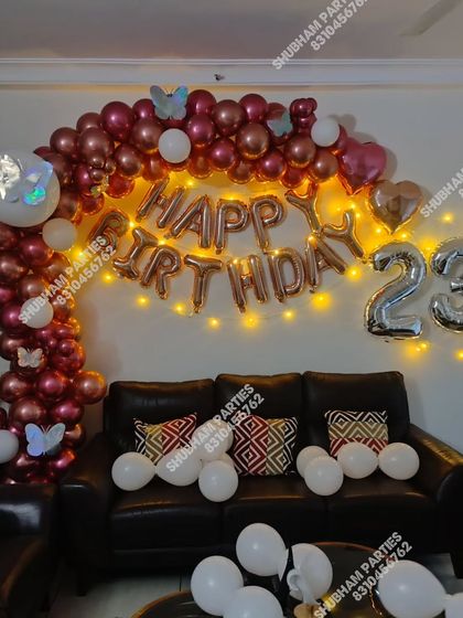 A stylish 23rd birthday setup in a living room, featuring a rose gold balloon arch over the sofa and fairy lights.
