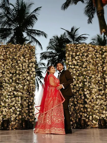 The happy couple posing in front of two massive floral walls made of white roses, set against a backdrop of palm trees and the evening sky.