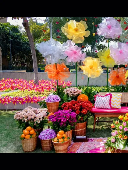A whimsical and colorful photo booth for a Mehendi party. The backdrop features giant, handcrafted paper flowers, while the foreground is filled with baskets of vibrant chrysanthemums and fresh oranges.