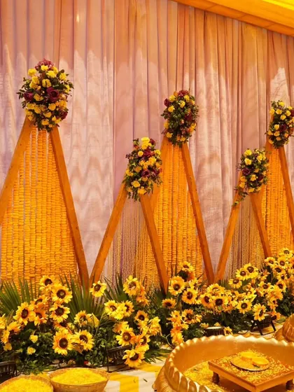 A unique Haldi backdrop design with triangular panels made of marigold strings. This setup includes a traditional urli and sunflower accents, perfect for an outdoor daytime event.