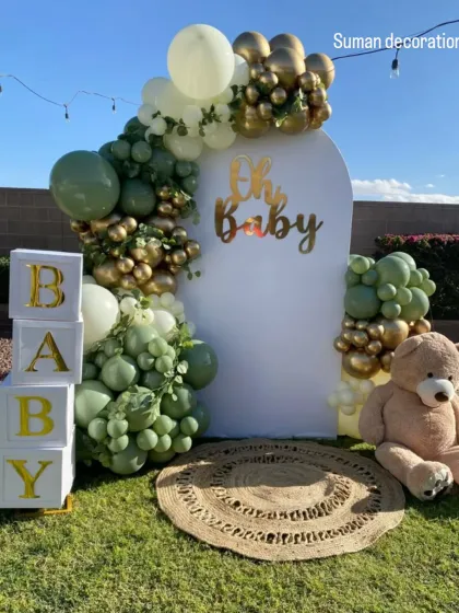 An outdoor "Oh Baby" shower with a rustic and natural theme. The decor includes a balloon arch in sage green, white, and gold, along with props like a teddy bear and "BABY" blocks.