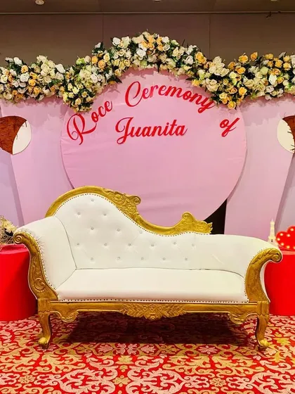 An elegant stage for a Roce ceremony, featuring a pink backdrop with a floral border and a classic white and gold chaise lounge. The design is both traditional and stylish.