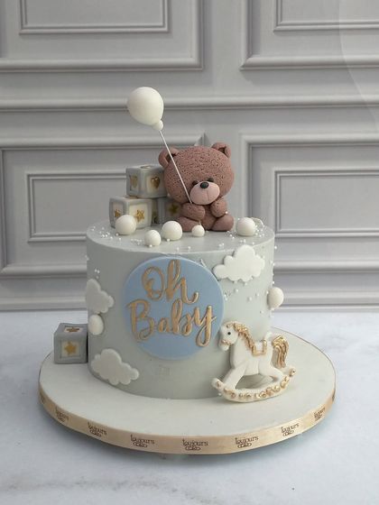 A sweet and gentle cake for a baby shower. The design features a soft blue color palette with clouds, a rocking horse, and an adorable teddy bear holding a balloon, with "Oh Baby" written in gold.