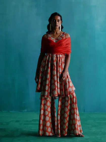 A contemporary ethnic wear look featuring a layered top and sharara pants. The styling with a bold red dupatta creates a striking color contrast against the blue background.