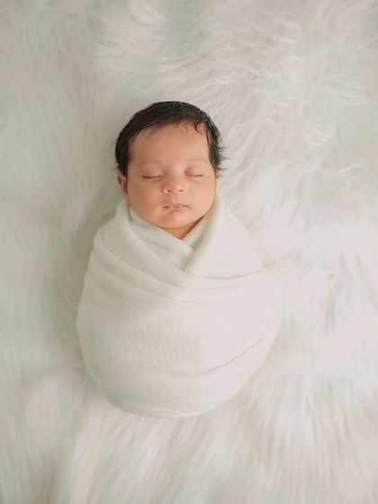 A simple and pure portrait of a newborn swaddled tightly in a white wrap, sleeping on a soft, fluffy white fur rug.