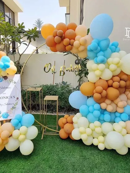 This outdoor baby shower decoration uses a gold ring backdrop and a vibrant balloon garland in orange, blue, and cream. A personalized welcome sign completes this cheerful and modern setup.