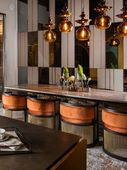 The warm, cheerful orange accents of the dining chairs create an inviting and energetic atmosphere. The reflective surfaces of the mirrored wall paneling and amber glass pendants enhance the ambient light.